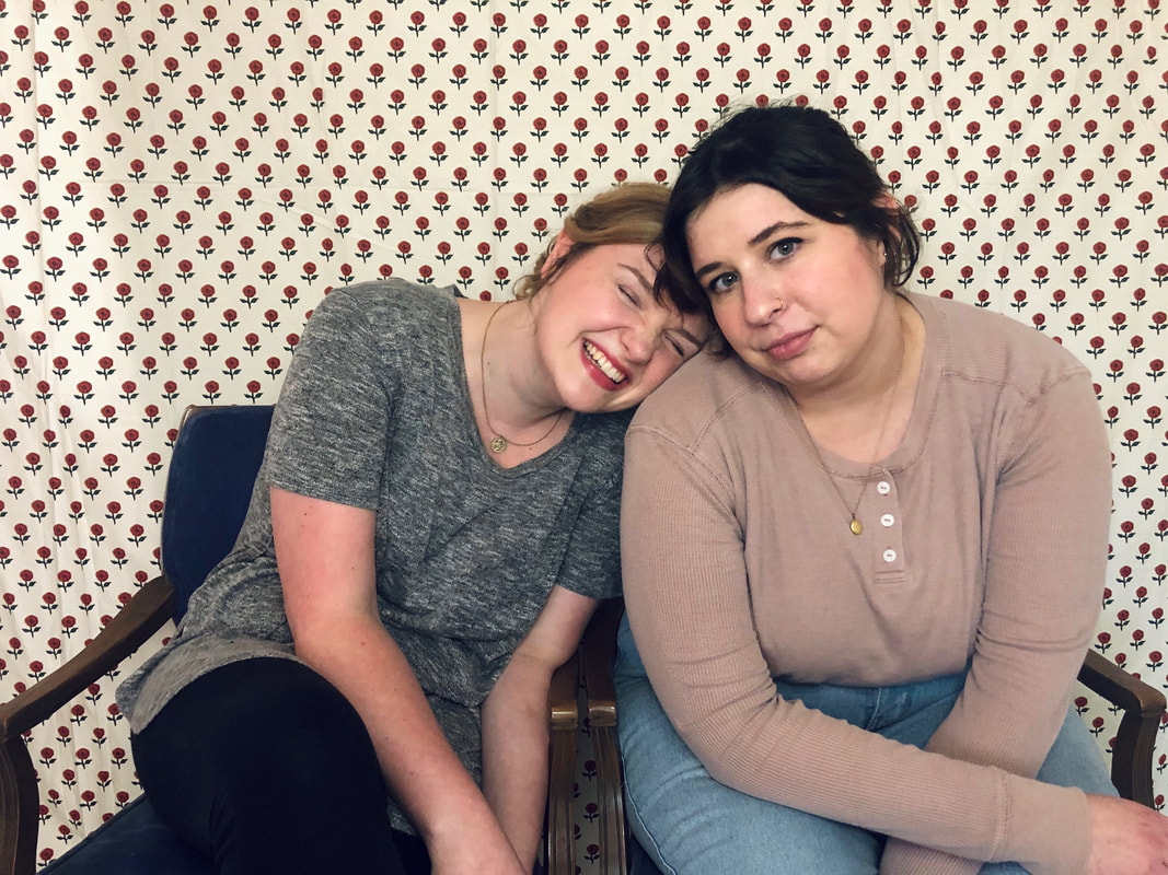 Image ID: A landscape photo of Jordan and Lex, sitting in blue velvet and wood chairs in front of a red and white floral background. Jordan, on the left, is wearing a grey shirt and leggings, and leans her head on Lex's shoulder while she laughs. Lex is wearing a light pink long sleeve shirt and light jeans, and sits with their arms crossed in front of them, looking at the camera with a neutral expression. End ID.