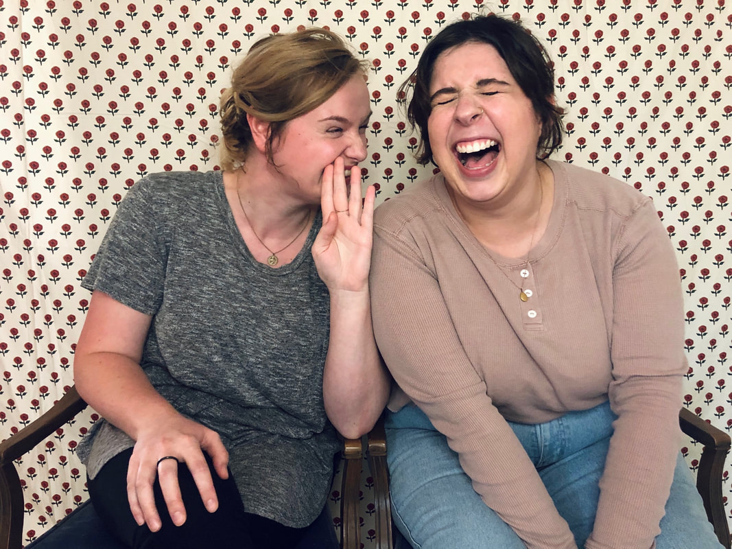 Image ID: Jordan and Lexare sitting next to each other in wood and blue velvet chairs, in front of a white and red floral backgroud. Jordan, wearing a grey shirt and leggings and a black ring on her right middle finger, is leaning in to Lex and laughing with one hand up to her mouth, as if telling a secret. Lex is wearing a light pink button up shirt and jeans, and has their hands clasped between their knees. They are leaning towards Jordan with their eyes shut and mouth open in laughter. End ID.