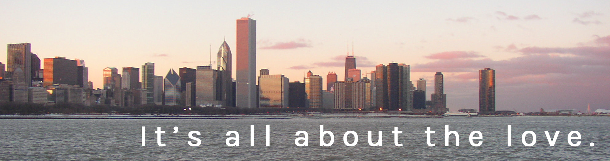 Image ID: A landscape photo of the Downtown Chicago skyline at sunset. In the foreground Lake Michigan is visible, and in the background, the sky is blue-grey and yellow, with pink clouds. In white sans-serif text, the bottom right corner of the image reads 