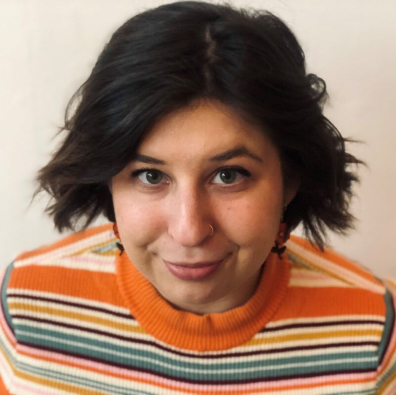 Image ID: A portrait photo of Lex, a white person with wavy, chin length dark brown hair. They are wearing an orange, white, yellow, blue, and purple striped sweater, and smiling mischeviously at the camera. End ID.
